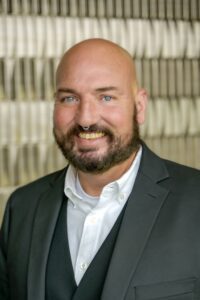 Portrait of a smiling man with a beard wearing a dark suit jacket and white shirt, standing in front of a softly blurred indoor background.