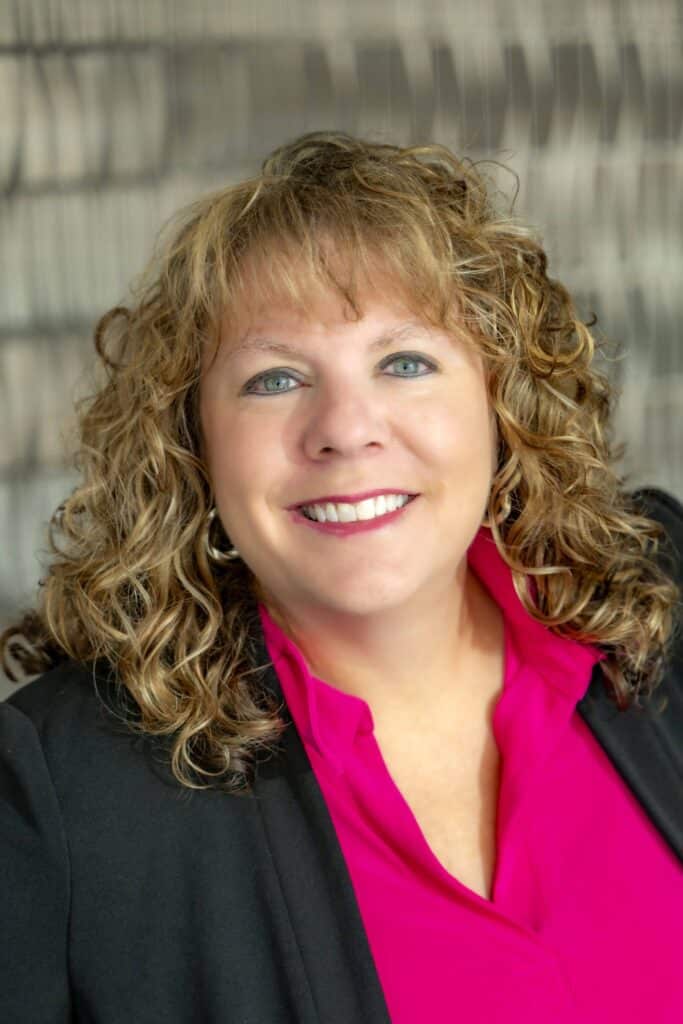 Professional headshot of a smiling woman with curly light brown hair wearing a bright pink blouse and black blazer, in front of a softly blurred neutral background.