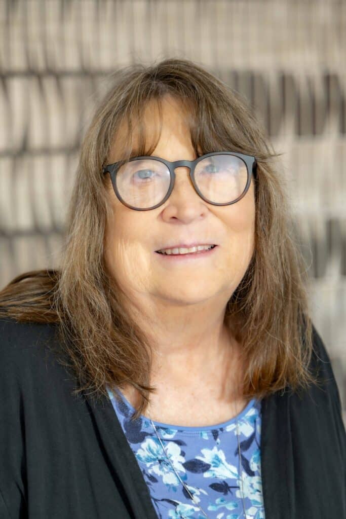 Professional headshot of a woman with shoulder-length brown hair wearing glasses, a black cardigan, and a blue floral blouse, in front of a softly blurred neutral background.