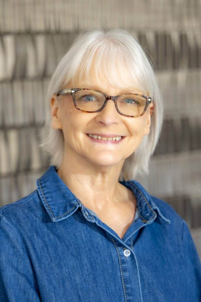 xProfessional headshot of a smiling woman with short light hair wearing glasses and a denim shirt, posed in front of a softly blurred neutral background.