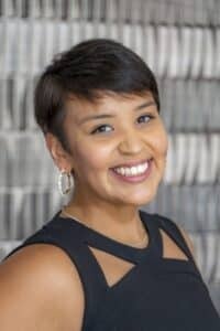 Professional headshot of a smiling woman with short dark hair wearing gold hoop earrings and a sleeveless black top, posed in front of a softly blurred neutral background.