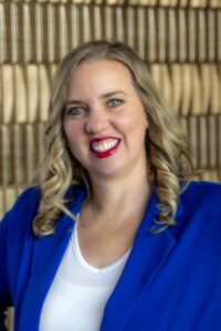 Smiling woman with shoulder-length blonde hair wearing a bright blue blazer and white top against a softly blurred neutral background.