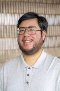 Portrait of a smiling man wearing glasses and a light gray collared shirt, standing in front of a softly blurred patterned background.