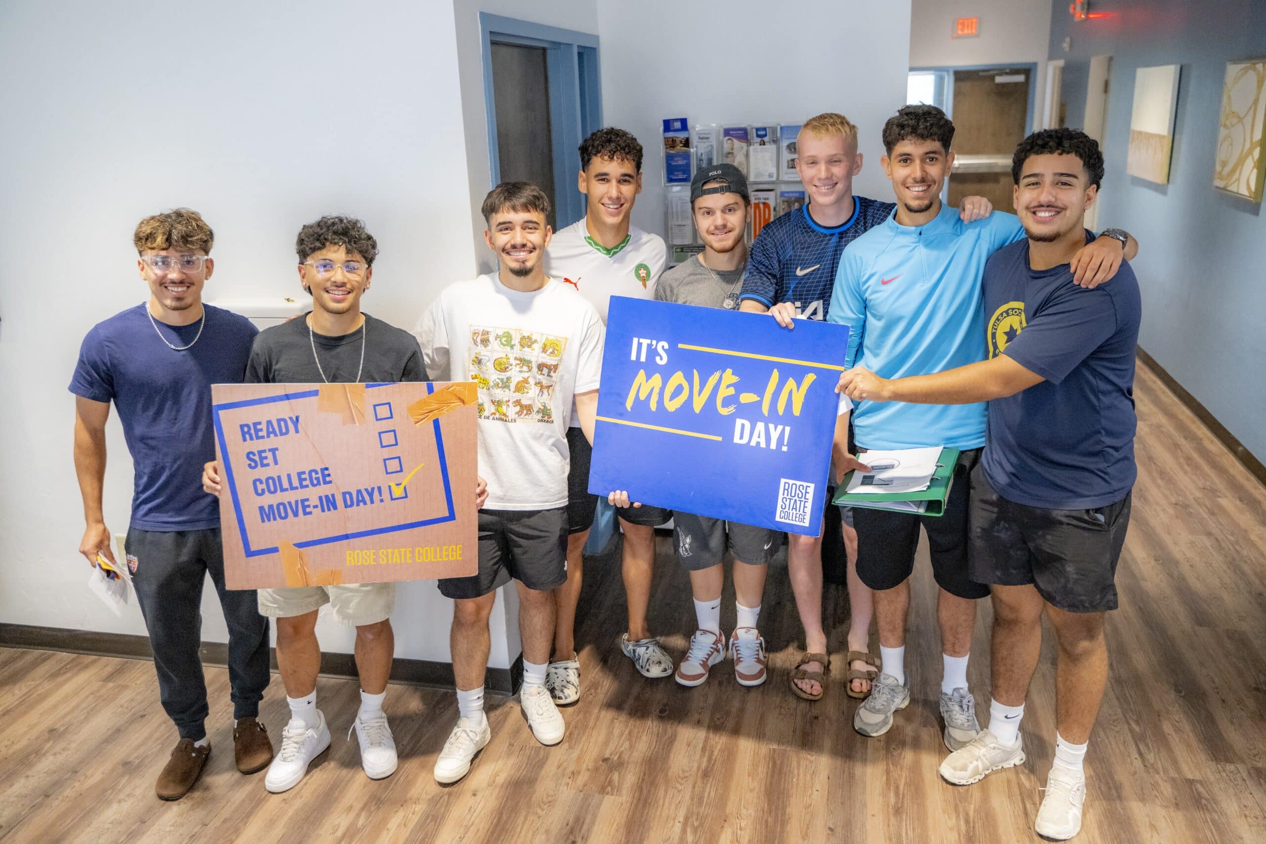 A group of male Rose State College students stand together in a hallway on move-in day, smiling and holding signs that read ‘Ready, Set, College, Move-In Day!’ and ‘It’s Move-In Day!'