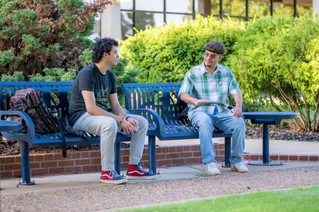 Two students sit on outdoor benches and talk with each other in a landscaped campus courtyard.