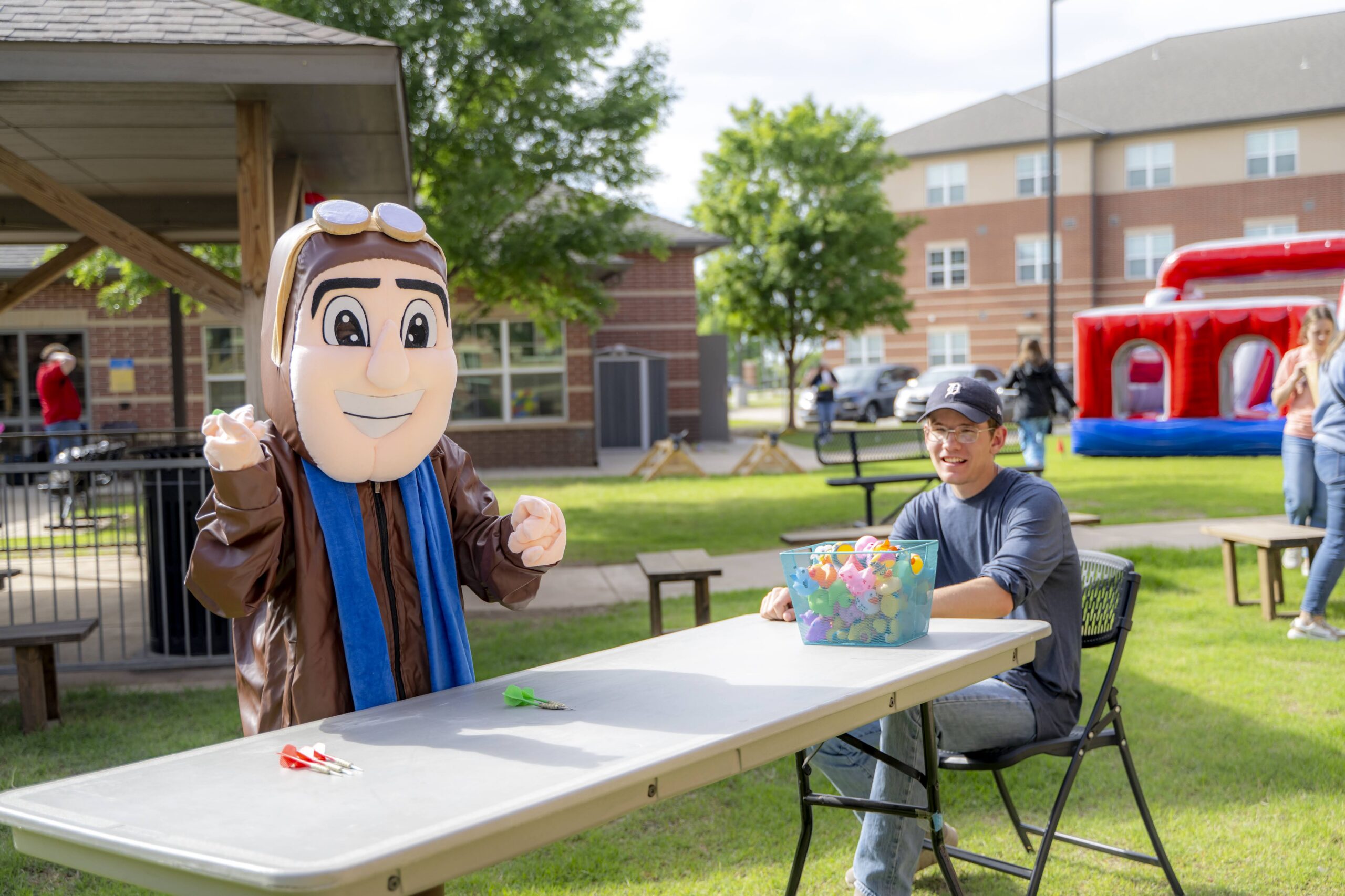 Rose State College’s Raider mascot stands beside a student volunteer at an outdoor carnival game table, with a basket of colorful plastic eggs and an inflatable activity area visible in the residence hall courtyard.