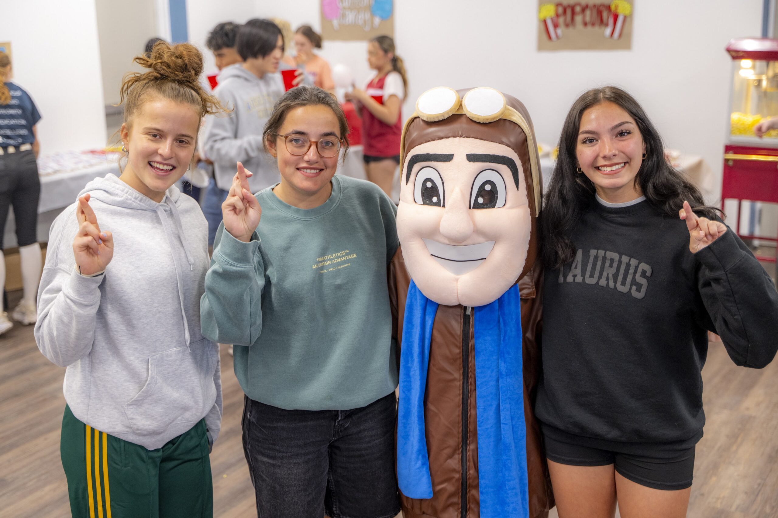 Three students pose with the Rose State College Raider mascot during an indoor residence hall carnival, smiling and holding up crossed fingers while other students participate in activities in the background.