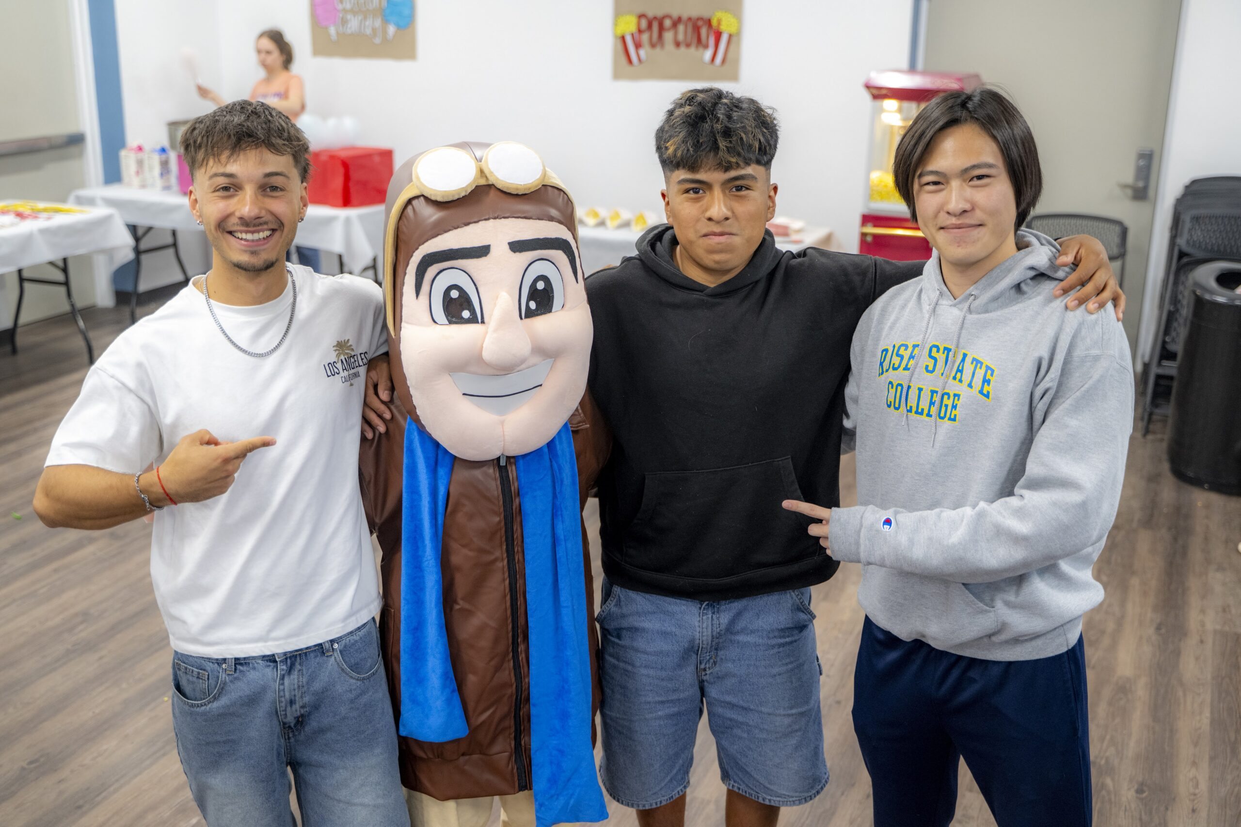 Three students pose with the Rose State College Raider mascot during an indoor residence hall carnival, smiling with their arms around each other as event activities and refreshments are visible in the background.