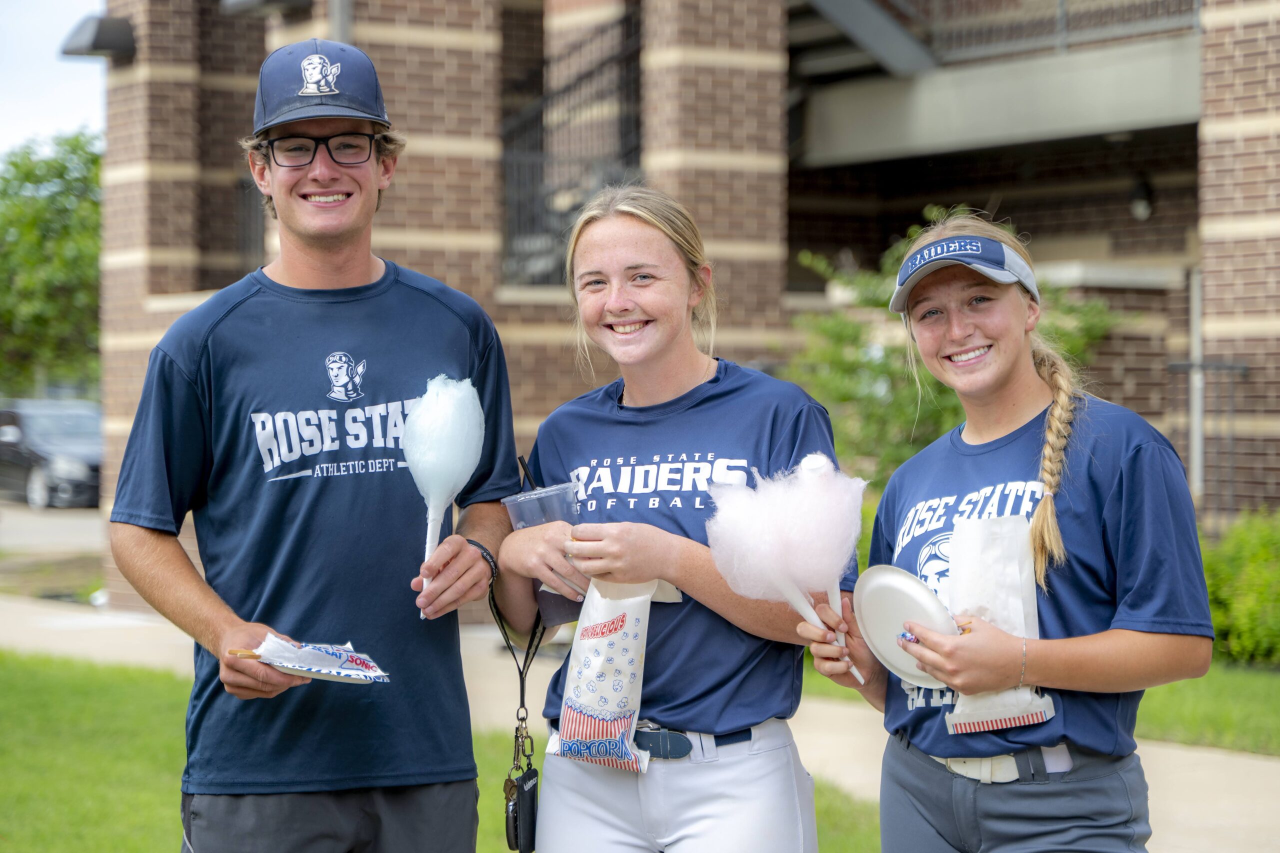 Three Rose State College student-athletes stand outdoors smiling during a residence hall carnival, holding cotton candy and snacks while wearing Raiders athletics apparel.