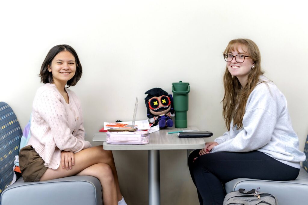 Two students sit across from each other at a study table, smiling toward the camera with notebooks, a laptop, and school supplies between them.