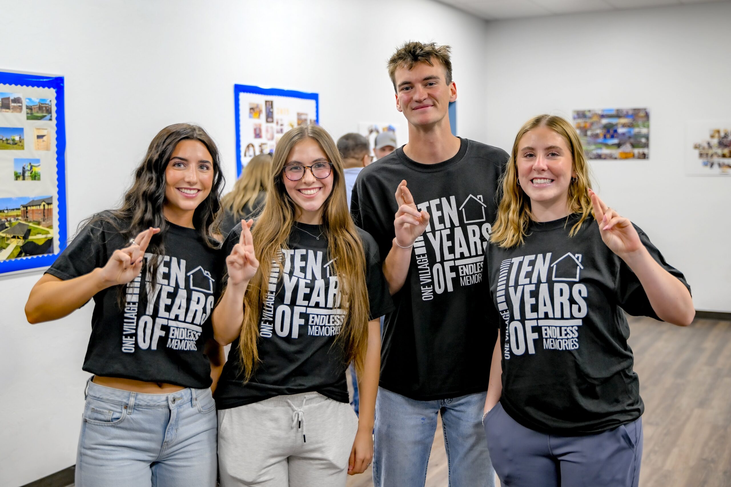 Four students wearing matching “Ten Years of Endless Memories” T-shirts smile and pose together during The Village’s 10th anniversary celebration, holding up crossed fingers in a community room decorated with photos and displays.