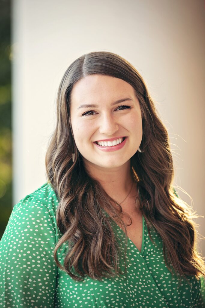 Professional headshot of a smiling woman with long brown hair wearing a green polka-dot blouse, photographed against a softly blurred background.