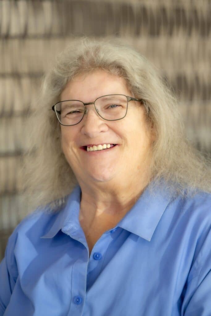 Professional headshot of a smiling woman with gray hair and glasses, wearing a light blue collared shirt, in front of a softly blurred neutral background.