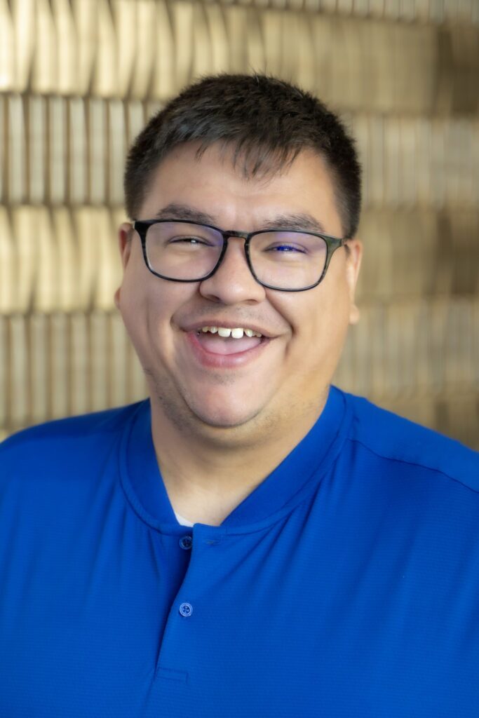 Smiling man with short dark hair and glasses wearing a blue shirt against a softly blurred neutral background.