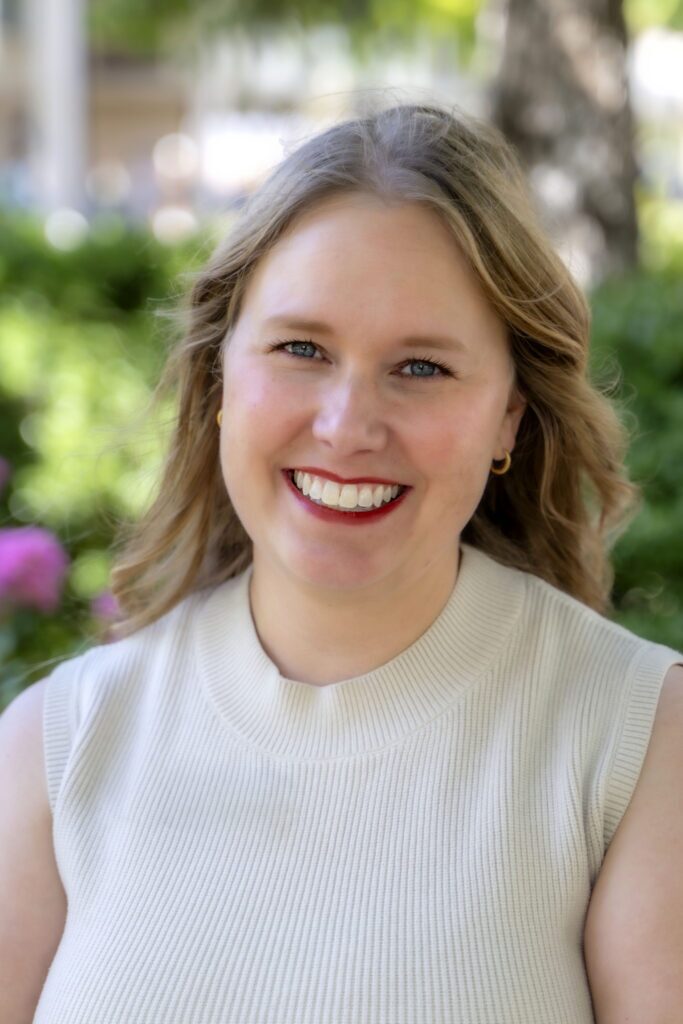 Professional headshot of a smiling woman with shoulder-length light brown hair wearing a sleeveless cream knit top, photographed outdoors with trees and greenery softly blurred in the background.