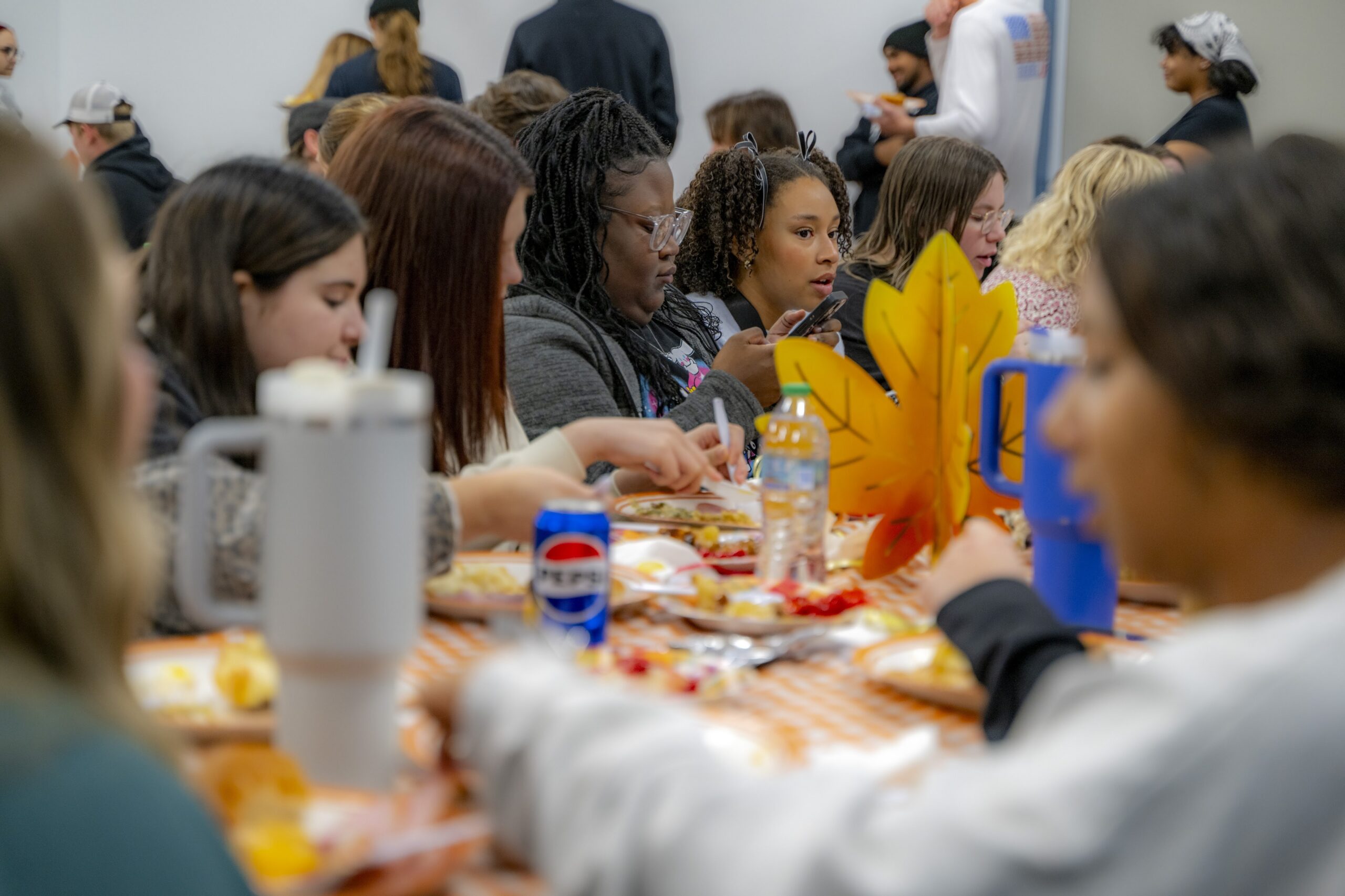 Students gather around a long table during a residence hall Thanksgiving event, sharing a meal and conversation in a decorated community room with autumn-themed table settings.