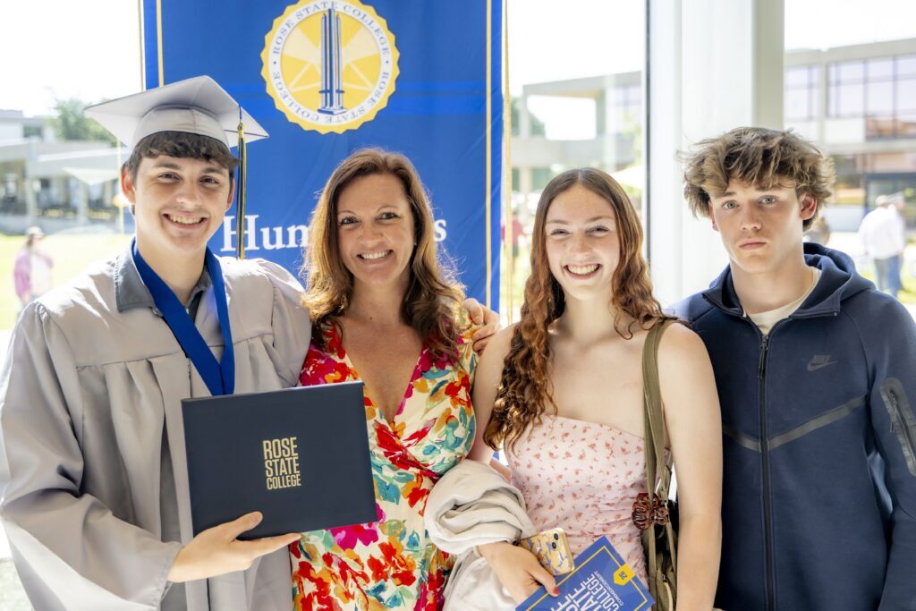 Rose State College graduate in a cap and gown holds a diploma cover while posing with three family members after commencement in the Student Union.