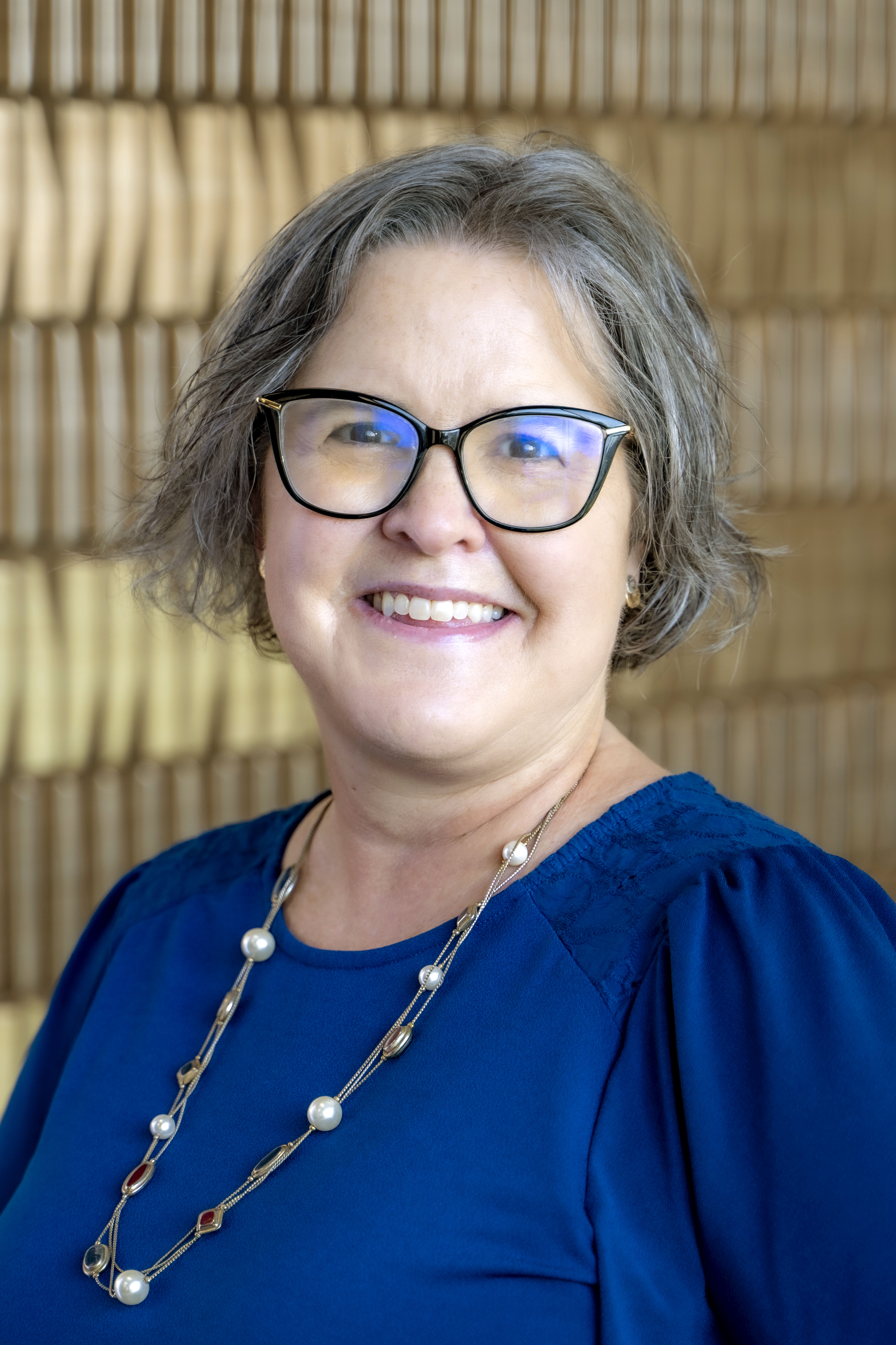 Smiling woman with short gray hair and glasses wearing a blue top and necklace against a softly blurred neutral background.
