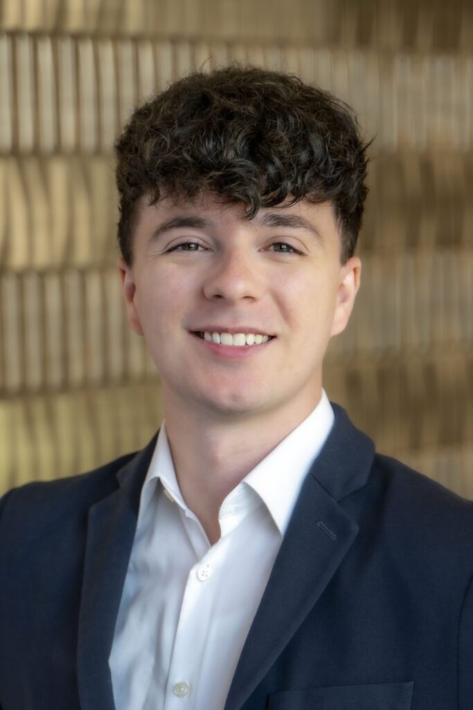 Smiling man with short curly dark hair wearing a navy blazer over a white collared shirt.