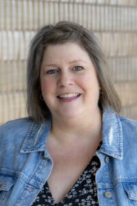 Professional headshot of a woman with shoulder-length brown hair wearing a denim jacket over a patterned blouse, smiling in front of a light brick wall background.