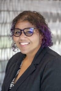 Professional headshot of a smiling woman wearing glasses and a black blazer, with curly brown and purple hair, standing in front of a light textured background.