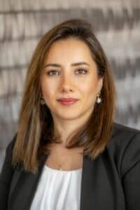 Professional headshot of a woman with straight brown hair, wearing a black blazer and white blouse, in front of a softly blurred neutral background.