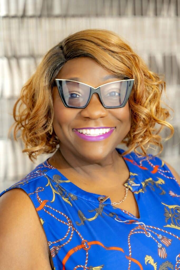 Professional headshot of a smiling woman with short curly hair and glasses, wearing a bright blue patterned blouse, in front of a softly blurred neutral background.