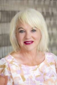 Professional headshot of a smiling woman with short blonde hair wearing a pastel floral blouse, in front of a softly blurred neutral background.
