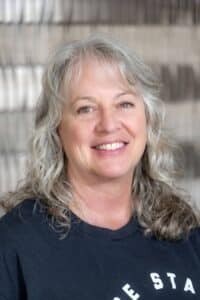 Professional headshot of a smiling woman with wavy gray hair wearing a black Rose State College shirt, in front of a softly blurred neutral background.