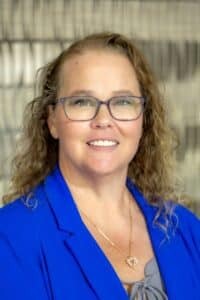 Professional headshot of a smiling woman with curly hair wearing glasses, a bright blue blazer, and a gold heart-shaped necklace, in front of a softly blurred neutral background.