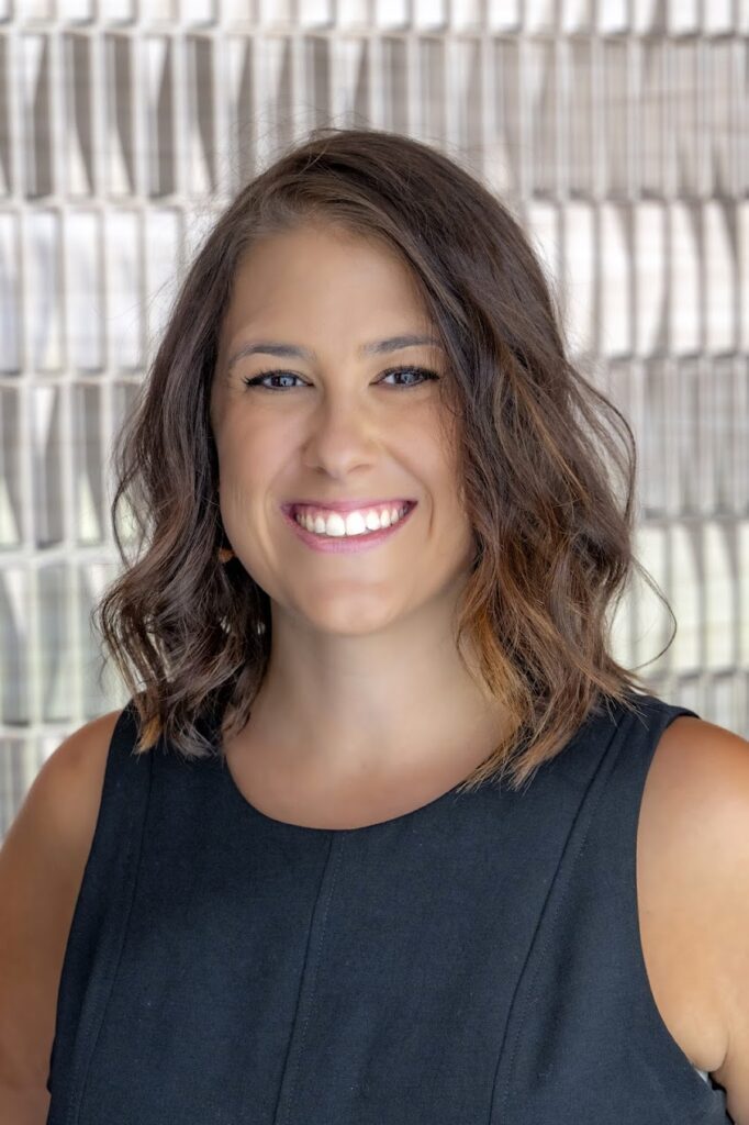 Portrait of a smiling woman with shoulder-length wavy brown hair, wearing a sleeveless black top, standing in front of a softly patterned background.