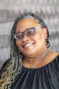 Professional headshot of a smiling woman with braided hair wearing glasses, hoop earrings, and a black blouse, in front of a softly blurred neutral background.