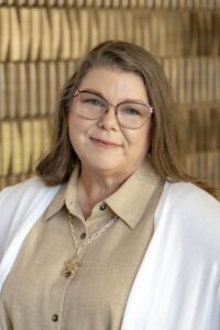 Smiling woman with shoulder-length brown hair and glasses wearing a tan collared shirt and white cardigan against a softly blurred neutral background.