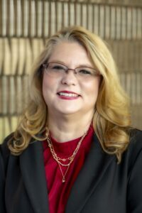 Christina Smith smiles for a professional headshot, wearing glasses, a red blouse, black blazer, and layered gold necklaces.