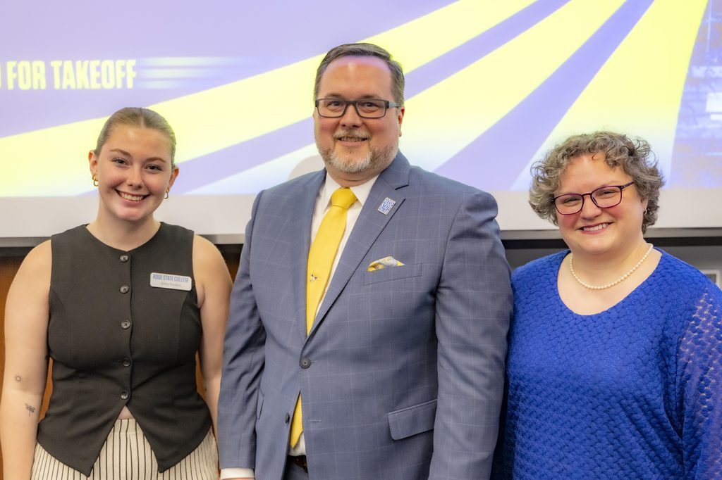 Three people smiling together at Rose State College’s State of Rose State event, with a man in a blue suit and yellow tie standing between two women in front of a Cleared for Takeoff presentation screen.