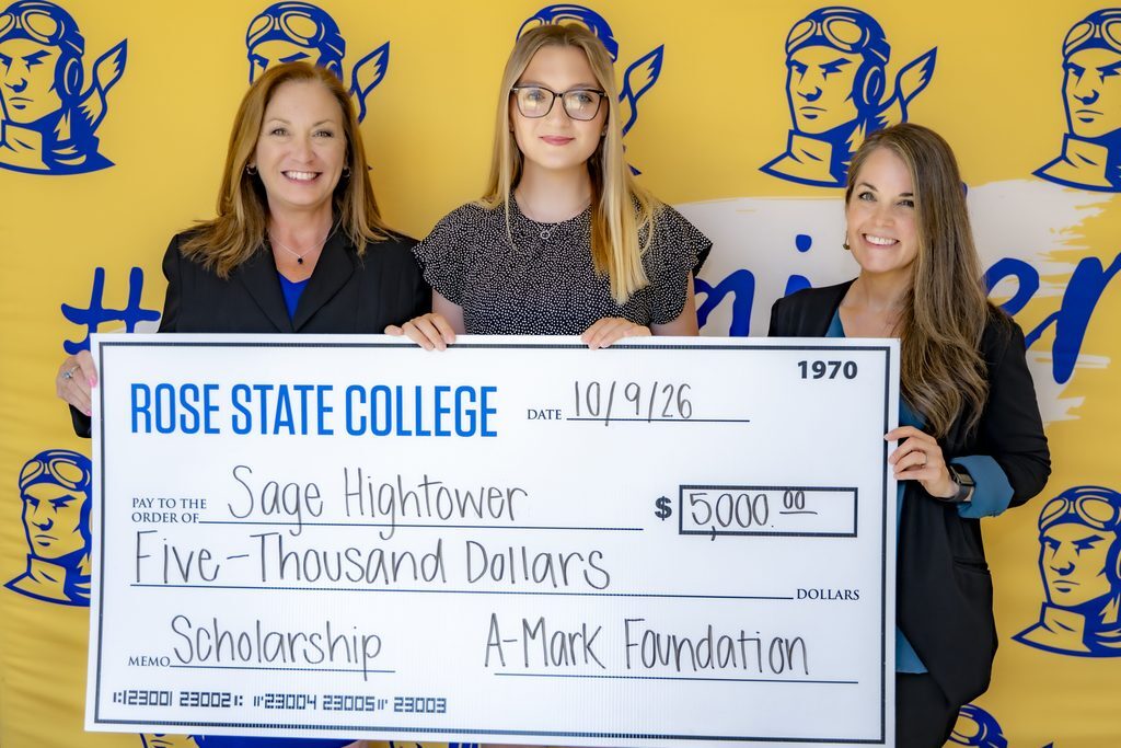 Rose State College student Sage Hightower stands with two women, holding a ceremonial $5,000 scholarship check from the A-Mark Foundation in front of a yellow and blue Rose State College backdrop.