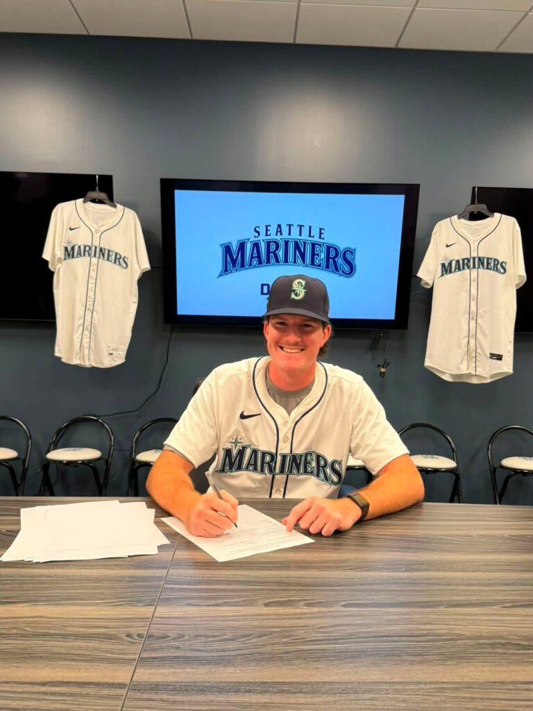 A student wearing a Seattle Mariners baseball uniform smiles while signing paperwork at a table beneath a display featuring the Seattle Mariners logo, with team jerseys hanging on the wall behind him.