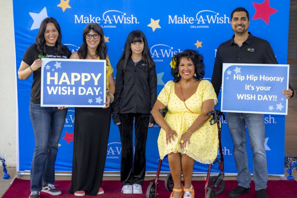 Five people pose in front of a Make-A-Wish Oklahoma backdrop, smiling and holding signs that read "Happy Wish Day" and "Hip Hip Hooray, it's your Wish Day!!" to celebrate a wish recipient.