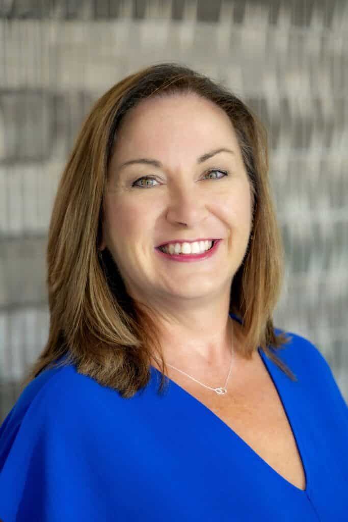 Professional headshot of a smiling woman with shoulder-length light brown hair, wearing a royal blue blouse and silver necklace, in front of a softly blurred neutral background.