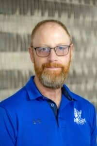 Professional headshot of a man with glasses and a beard, wearing a blue Rose State College polo shirt, in front of a softly blurred neutral background.