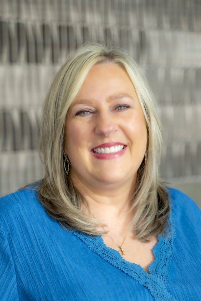 Professional headshot of a smiling woman with blonde hair wearing a blue blouse, in front of a softly blurred neutral background.