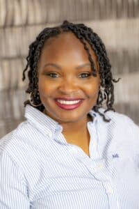 Professional headshot of a smiling woman with short twisted hair wearing hoop earrings and a light blue striped button-down shirt, in front of a softly blurred neutral background.