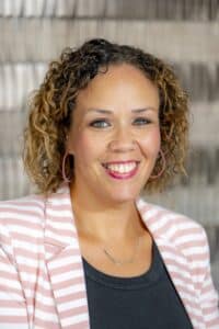 Professional headshot of a smiling man with glasses and a short beard wearing a light blue button-up shirt, in front of a softly blurred neutral background.