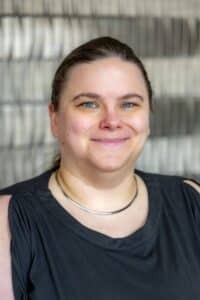 Professional headshot of a smiling woman with pulled-back hair wearing a sleeveless black top and a silver necklace, in front of a softly blurred neutral background.