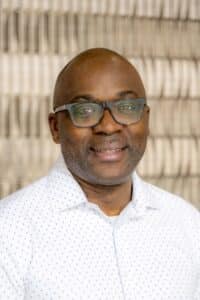 Professional headshot of a smiling man wearing glasses and a white dress shirt with a subtle pattern, in front of a softly blurred neutral background.