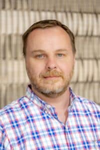 Professional headshot of a man with short hair and a beard wearing a red, white, and blue plaid shirt, in front of a softly blurred neutral background.