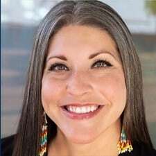 Portrait of a smiling woman with dark hair wearing multi-colored earrings and a black top photographed against a light textured background.