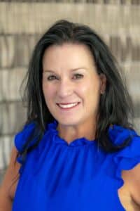 Professional headshot of a smiling woman with long dark hair, wearing a bright blue blouse, in front of a softly blurred neutral background.