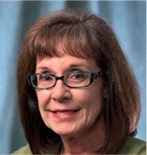 Professional headshot of a smiling woman with shoulder-length brown hair and glasses, wearing a green top, in front of a light blue background.