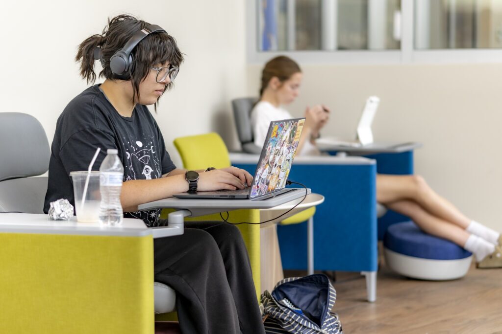 A student wearing headphones works on a laptop in a campus lounge while another student studies in the background.